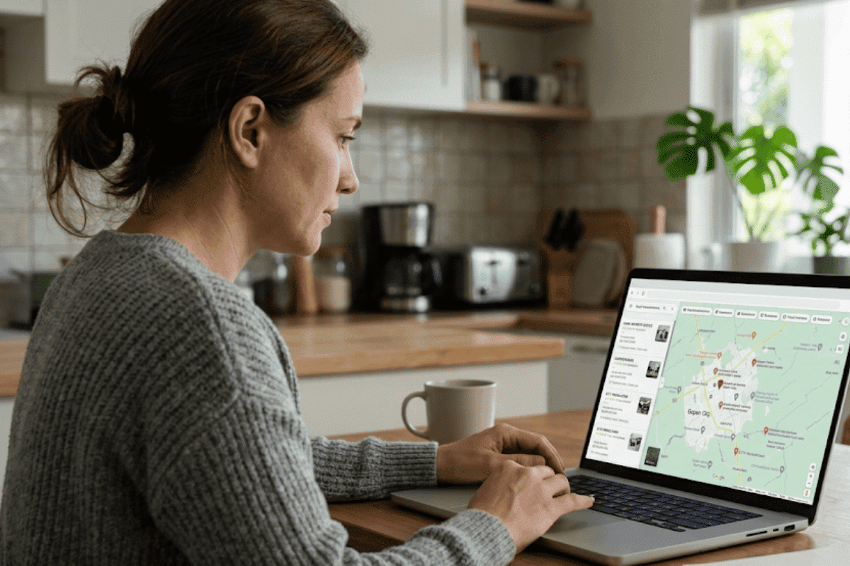 Woman in a gray sweater sits at a kitchen table typing on a laptop with a map and pins displayed on the screen.
